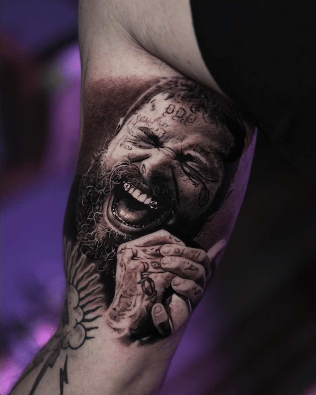 Tattoo of a smiling man with a beard and facial tattoos, showcasing intricate details and shading, on an arm with additional tattoo designs, emphasizing tattoo artistry and personal expression.